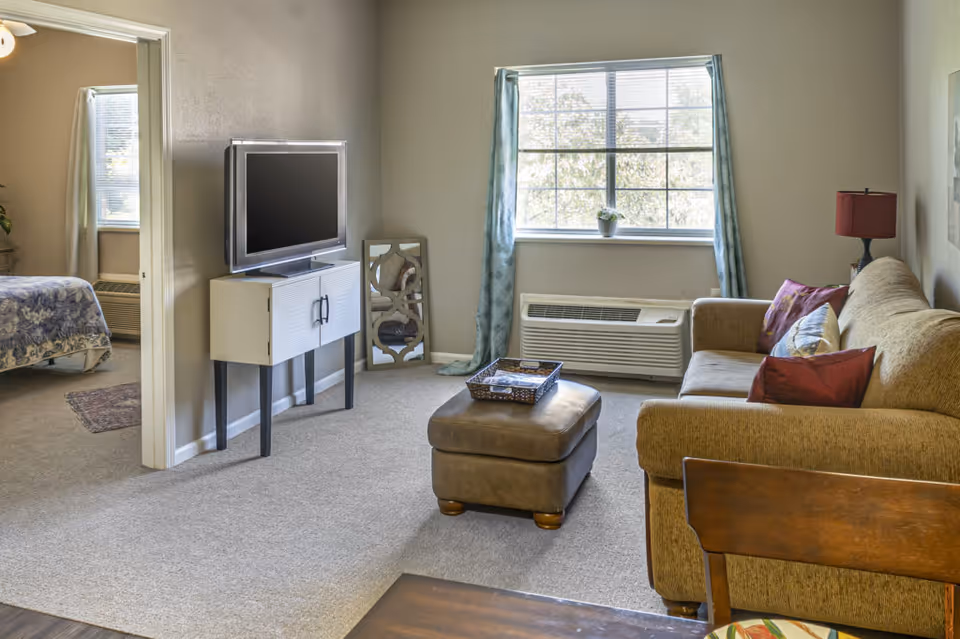 A cozy living room area with a beige sofa adorned with colorful pillows, a brown ottoman with a decorative tray on top, a flat-screen TV on a white stand, and a window with blue curtains letting in natural light. In the background, a bedroom with a bed covered in a blue floral bedspread is visible through an open doorway.