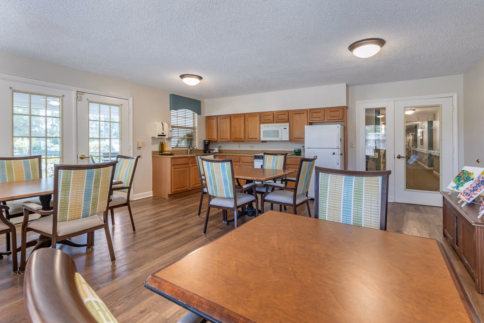 Bright communal dining area with several tables and chairs and an open kitchen with wooden cabinets and appliances.