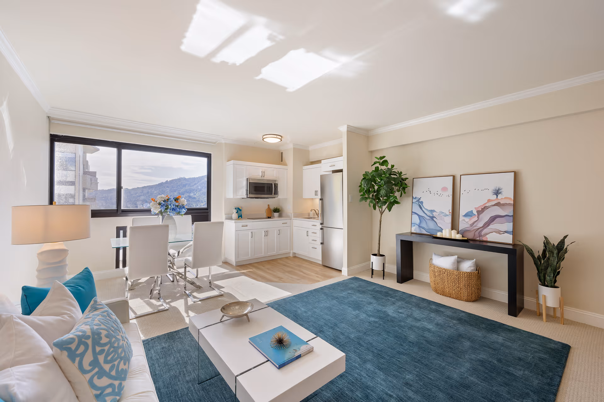 Bright open-plan living room with a white sofa, blue rug, glass dining table, small kitchenette, large window and wall artwork.