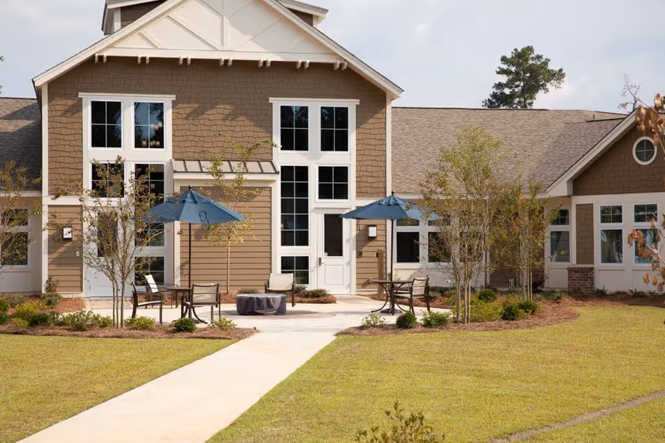 Front exterior of a senior living building with patio seating, umbrellas, a central walkway, and landscaped lawn.