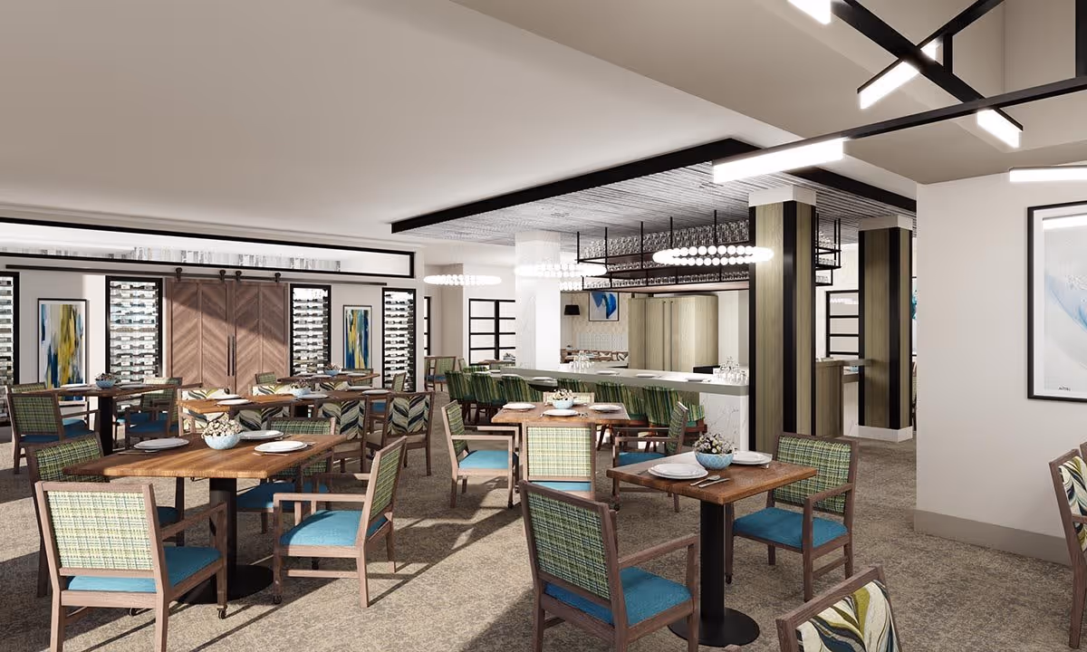 A spacious dining room with multiple wooden tables and chairs featuring green and blue upholstery. The tables are set with plates, bowls, and cutlery. The room has modern lighting fixtures, large windows, and decorative artwork on the walls. There is a bar area with high chairs and glassware hanging above it.