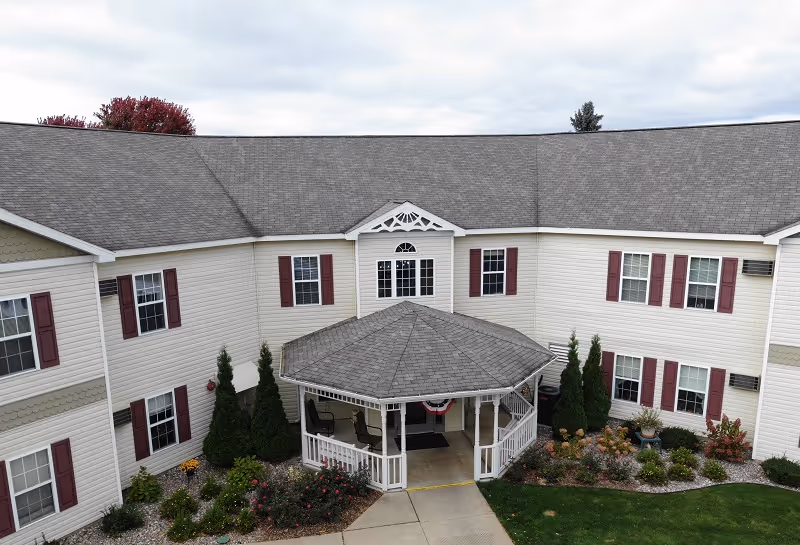 Exterior view of a two-story senior living facility building with beige siding and maroon shutters. The building has a gray shingled roof and a covered entrance with a small porch and white railing. There are landscaped bushes and flowers around the entrance and a concrete walkway leading to the door. The sky is cloudy.