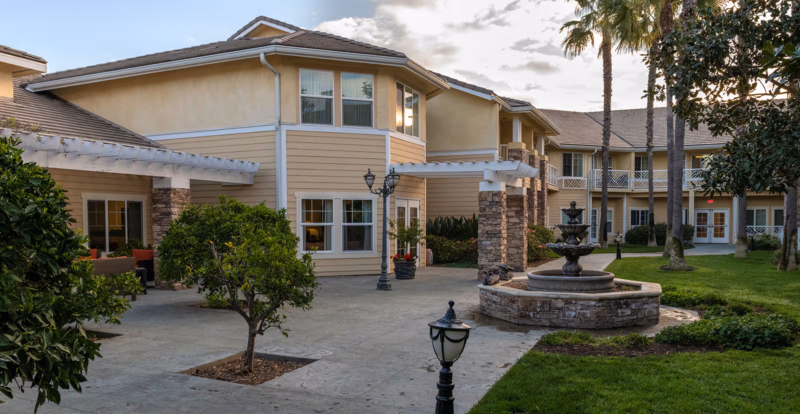 Outdoor courtyard area of a senior living facility with a multi-tiered stone fountain, green grass, palm trees, and a beige two-story building with white trim and balconies in the background.