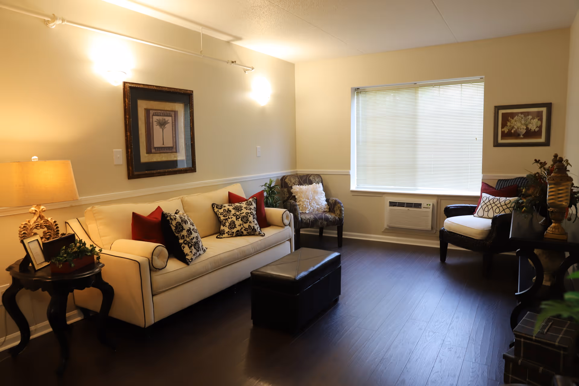 Well-lit living room with a cream sofa, patterned chairs, decorative pillows, side tables, and a window with blinds.