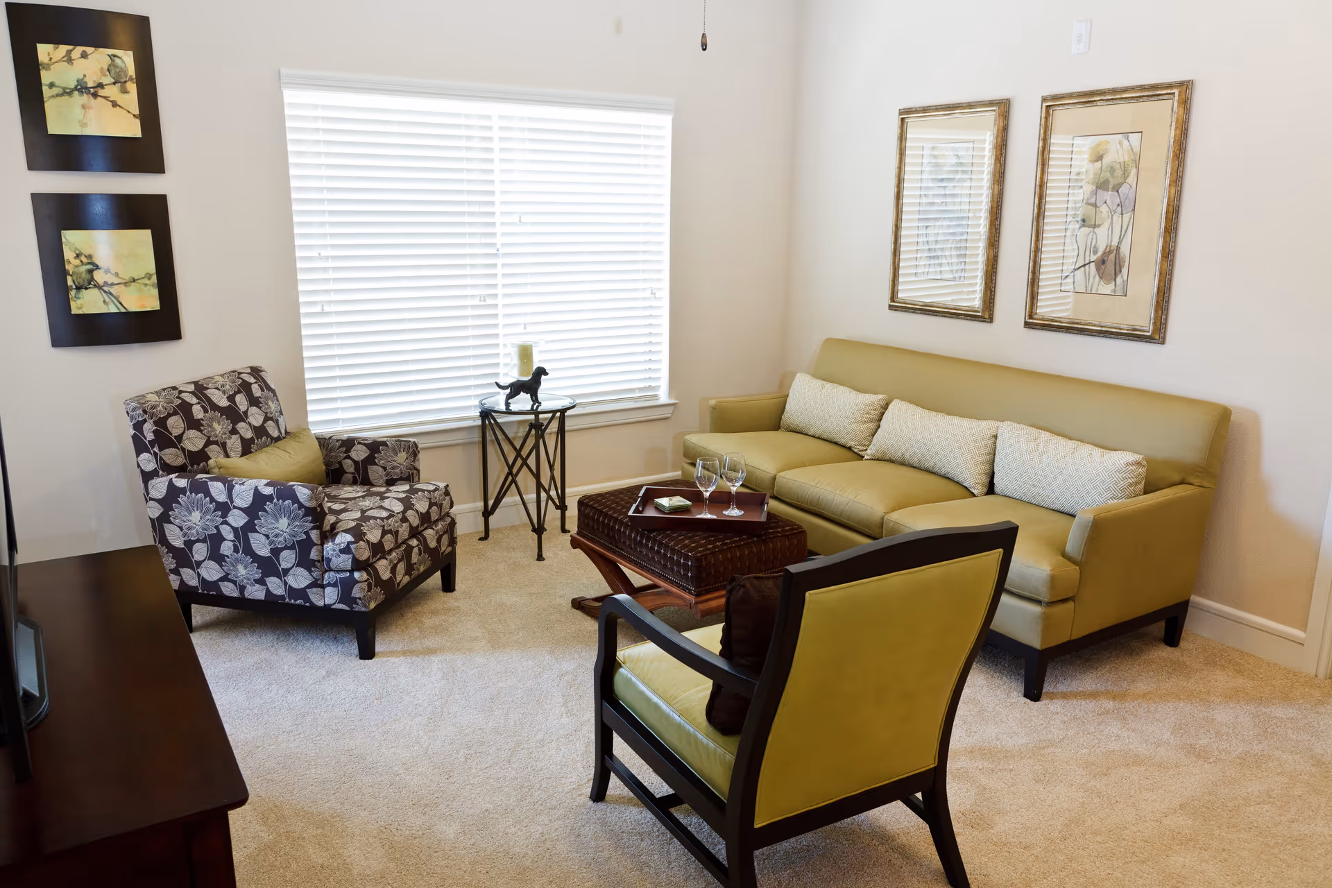 A cozy living room with a green sofa, two armchairs—one with floral upholstery and the other matching the sofa—and a small wooden coffee table with two wine glasses and a tray. The room has beige carpeting, a large window with white blinds, two framed artworks on the wall above the sofa, and two smaller framed pictures on the adjacent wall.