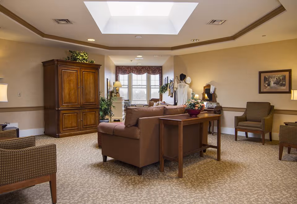 A cozy living room area with beige walls and carpet, featuring a brown sofa, two patterned armchairs, a wooden cabinet, a wooden table with a flower arrangement, and a large skylight in the ceiling. The room is softly lit with lamps and natural light from a window with decorative curtains.