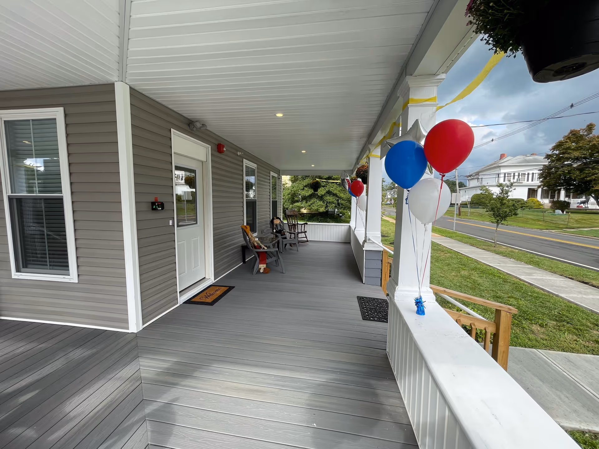 A wide covered porch of a residential building with gray siding and white trim. The porch has several chairs, a welcome mat in front of a white door, and red, white, and blue balloons tied to the white railing. The porch overlooks a sidewalk and a street with houses and trees in the background under a cloudy sky.