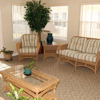 Sunlit sitting area with wicker sofa and armchair, glass-top coffee table, potted plants, and a lamp in front of bay windows.