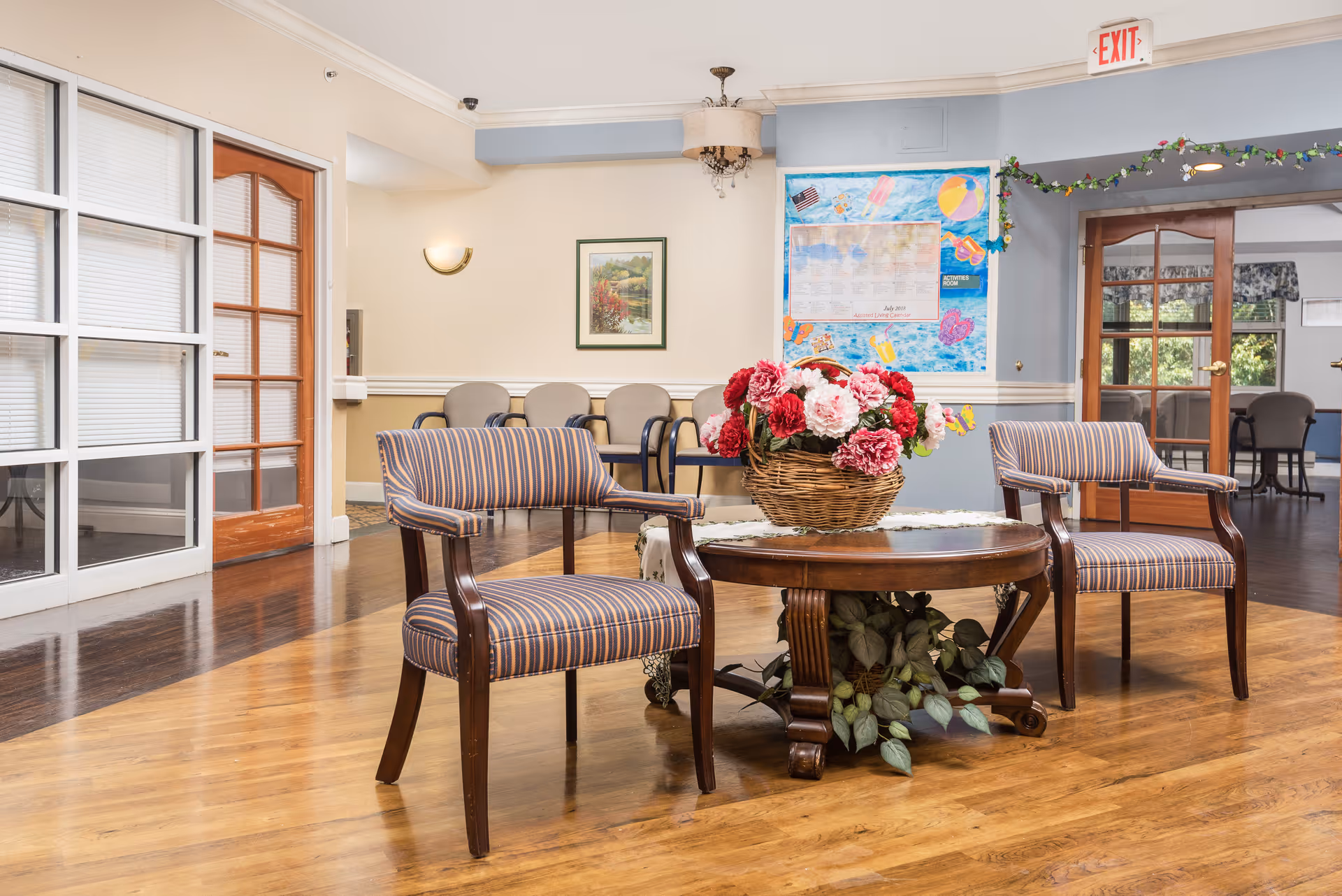 A seating area in a senior living facility with two striped armchairs around a wooden round table that has a basket of red and pink flowers on it. In the background, there are additional chairs lined up against the wall, a framed painting, a colorful bulletin board, and double wooden doors leading to another room.