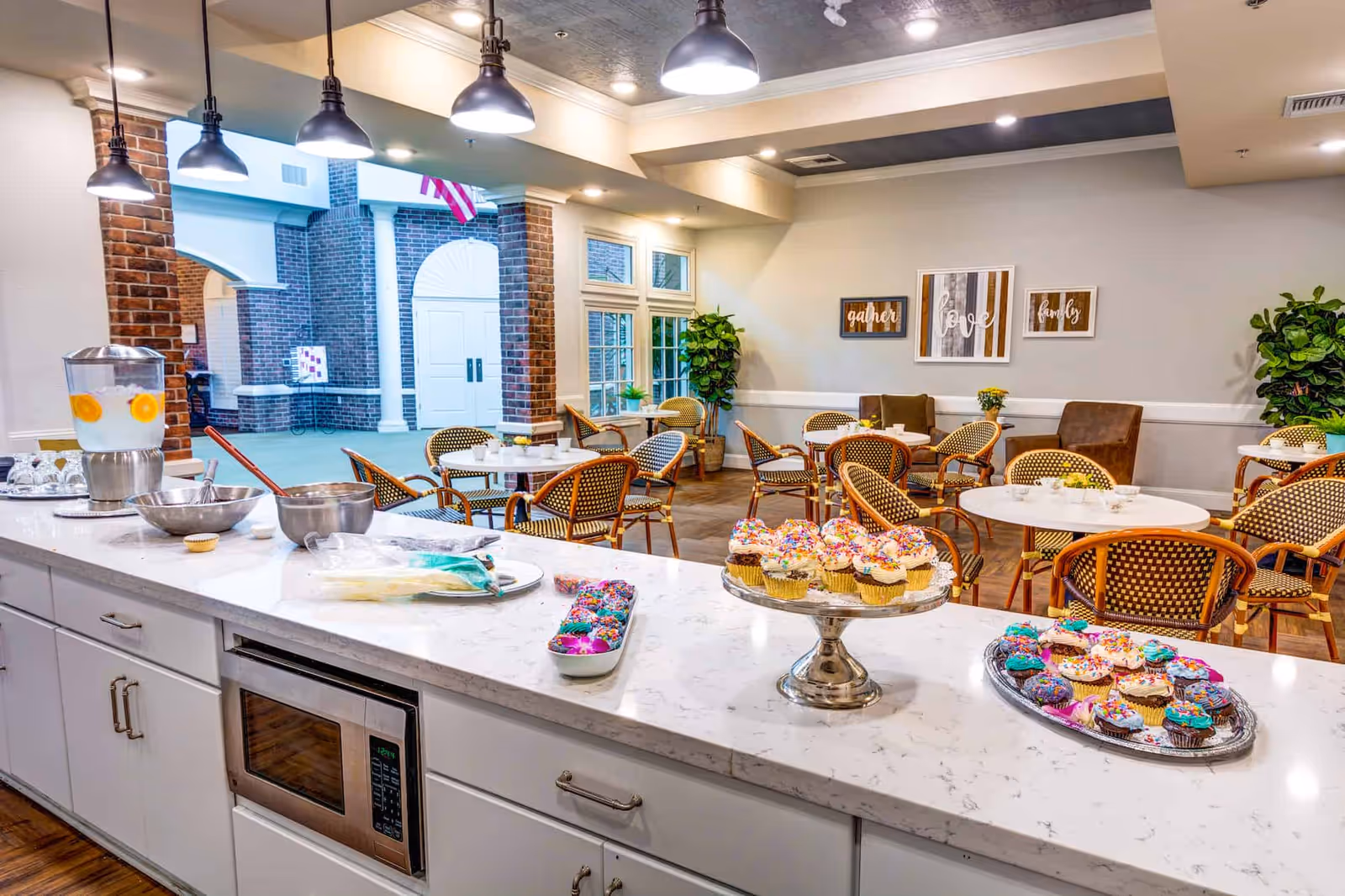 Bright communal dining area with a marble countertop buffet displaying cupcakes and a beverage dispenser, round tables and wicker chairs under pendant lights.