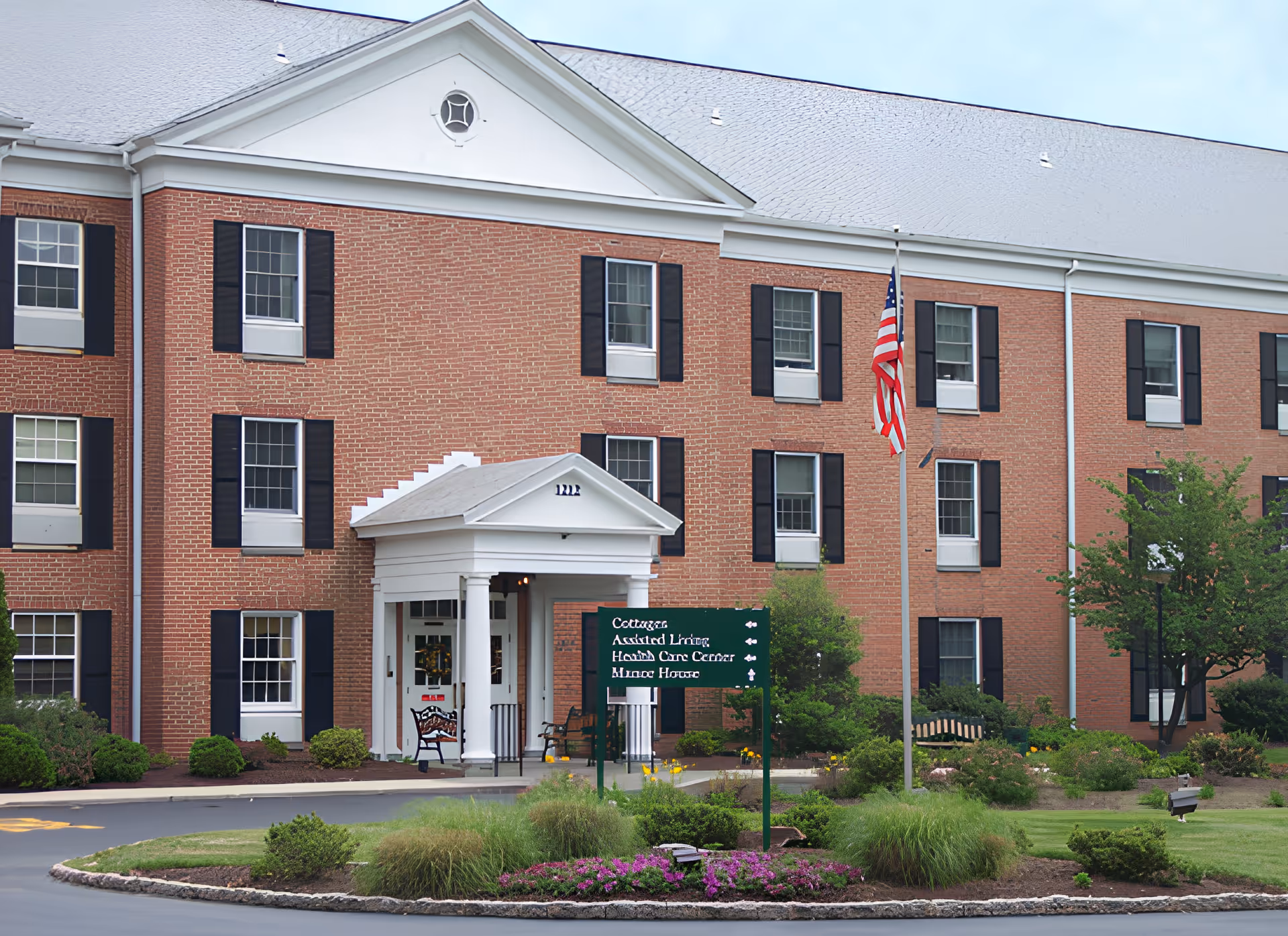 Exterior view of a large brick senior living facility building with white trim and black shutters. The entrance features a white portico with columns. There is a green sign in front indicating directions to cottages, assisted living, health care center, and manor house. An American flag is flying on a flagpole near the entrance, and there are landscaped bushes and flowers around the driveway.