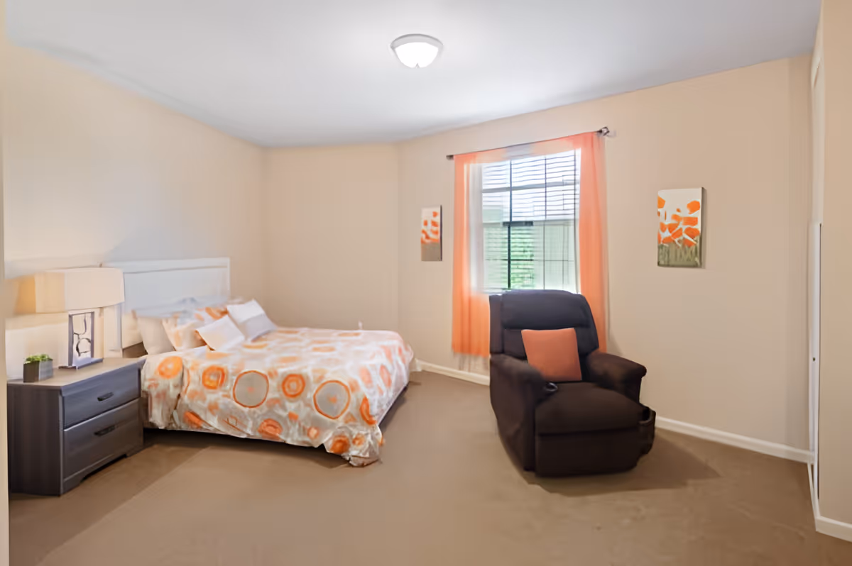 A bedroom with a bed covered in a white and orange patterned blanket, a dark wooden nightstand with a lamp and small plant, a large window with orange curtains, a dark brown recliner chair with an orange pillow, and two paintings with orange floral designs on the beige walls.