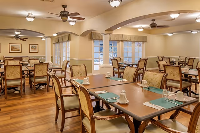 A spacious dining room with multiple wooden tables and chairs arranged neatly. Each table is set with placemats, napkins, cups, and glasses. The room has warm lighting, ceiling fans, and large windows with curtains allowing natural light to enter. The floor is wooden, and the walls are painted in a soft beige color.