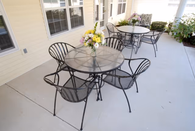 Outdoor patio area with two round metal tables, each surrounded by four metal chairs. Each table has a vase with a bouquet of flowers. The patio is adjacent to a building with cream-colored siding and multiple windows and doors.