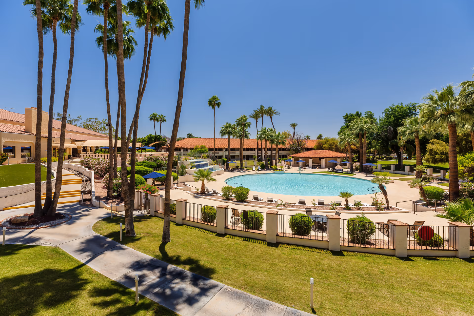 Outdoor view of a senior living facility featuring a large circular swimming pool surrounded by lounge chairs and umbrellas. Tall palm trees and well-maintained green lawns are visible around the pool area, with buildings in the background under a clear blue sky.