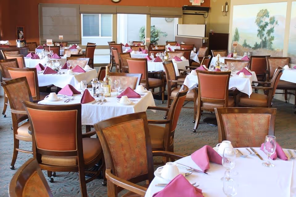 A dining room with multiple tables covered in white tablecloths, each set with purple folded napkins, glassware, cups, and silverware. The room has wooden chairs with cushioned seats and backs, large windows letting in natural light, and a painting on the wall.
