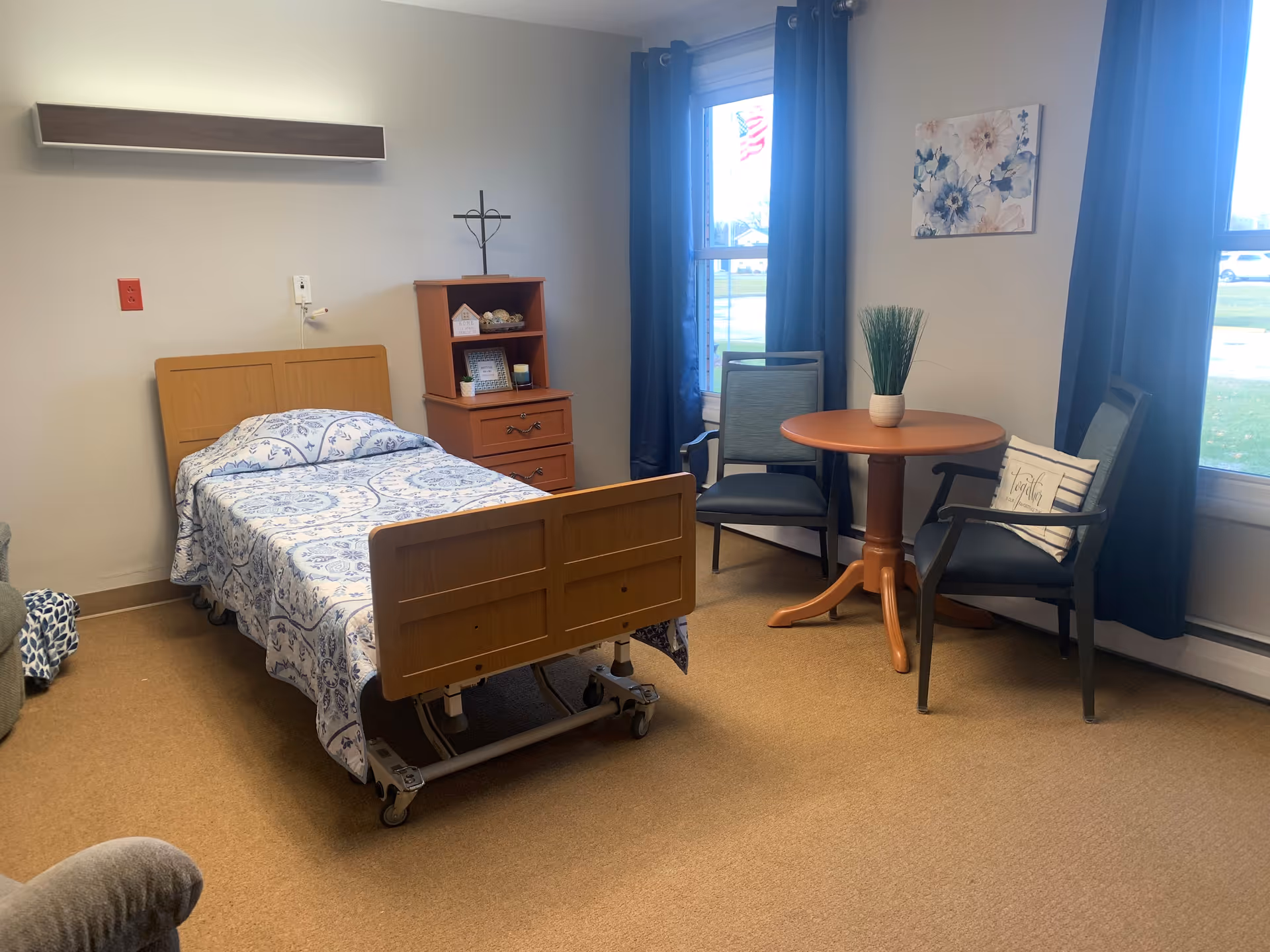 A furnished senior living bedroom with an adjustable single bed, bedside cabinet and shelving, and a small round table with two chairs by the windows.