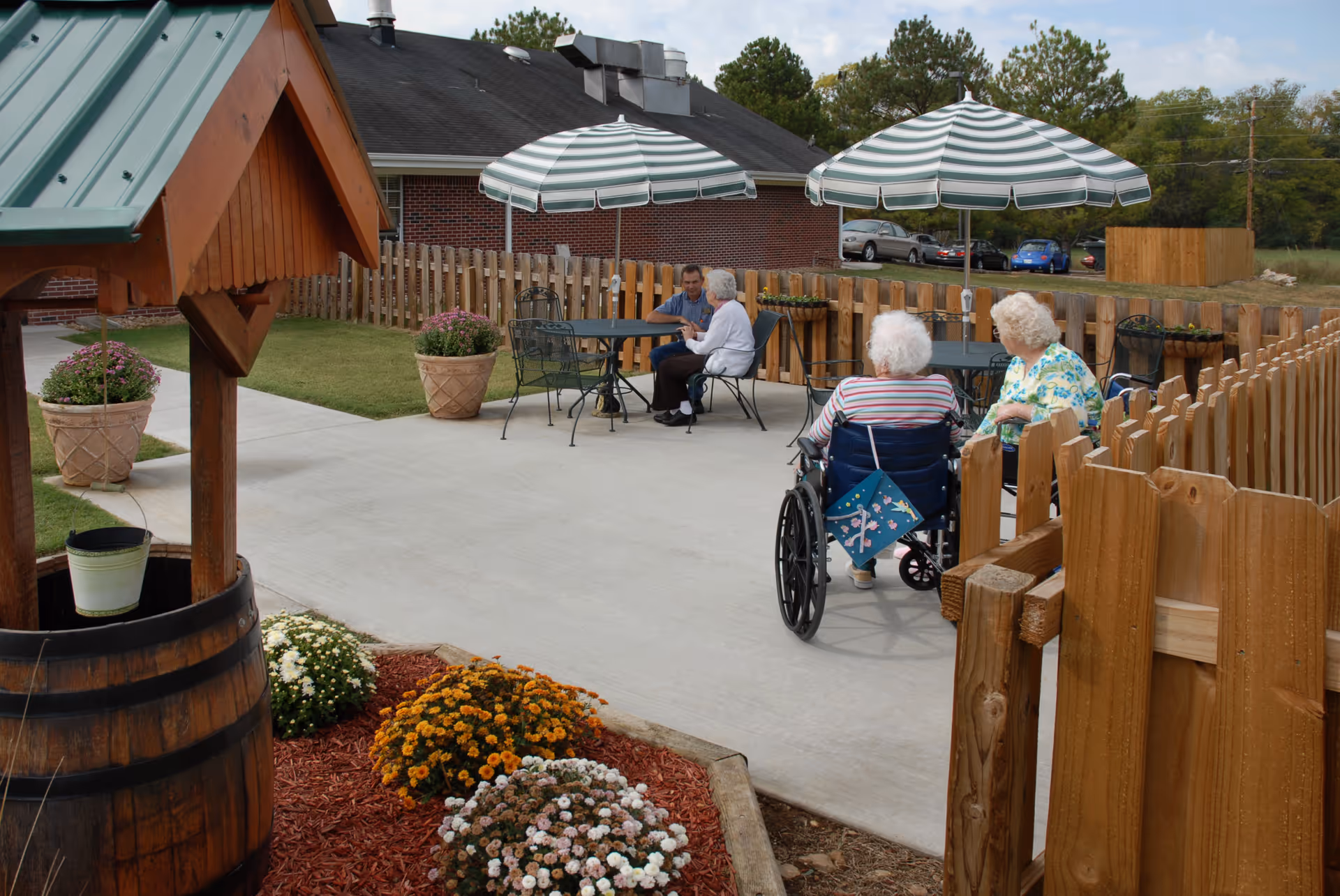 Outdoor patio area at Lynchburg Nursing Center with elderly residents sitting at tables under striped umbrellas. The area is enclosed by a wooden fence and features potted plants and a decorative wooden wishing well near flower beds.