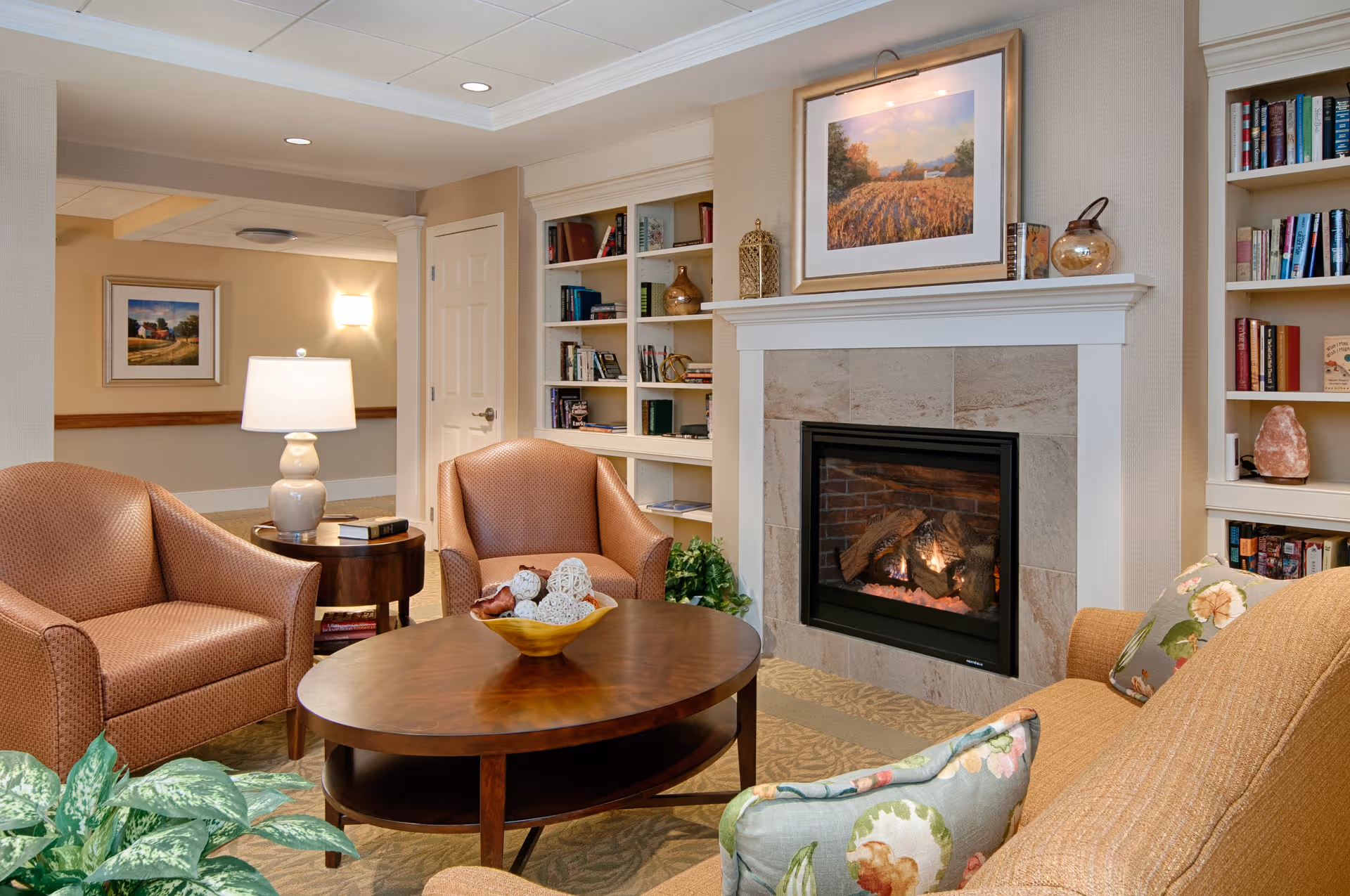 A cozy living room area in Golden View Health Care Center featuring a lit fireplace with a framed landscape painting above it. The room has two brown armchairs, a wooden coffee table with decorative items, a side table with a lamp and books, built-in bookshelves filled with books and decorative objects, and a sofa with floral pillows. The walls are beige and the floor is carpeted.