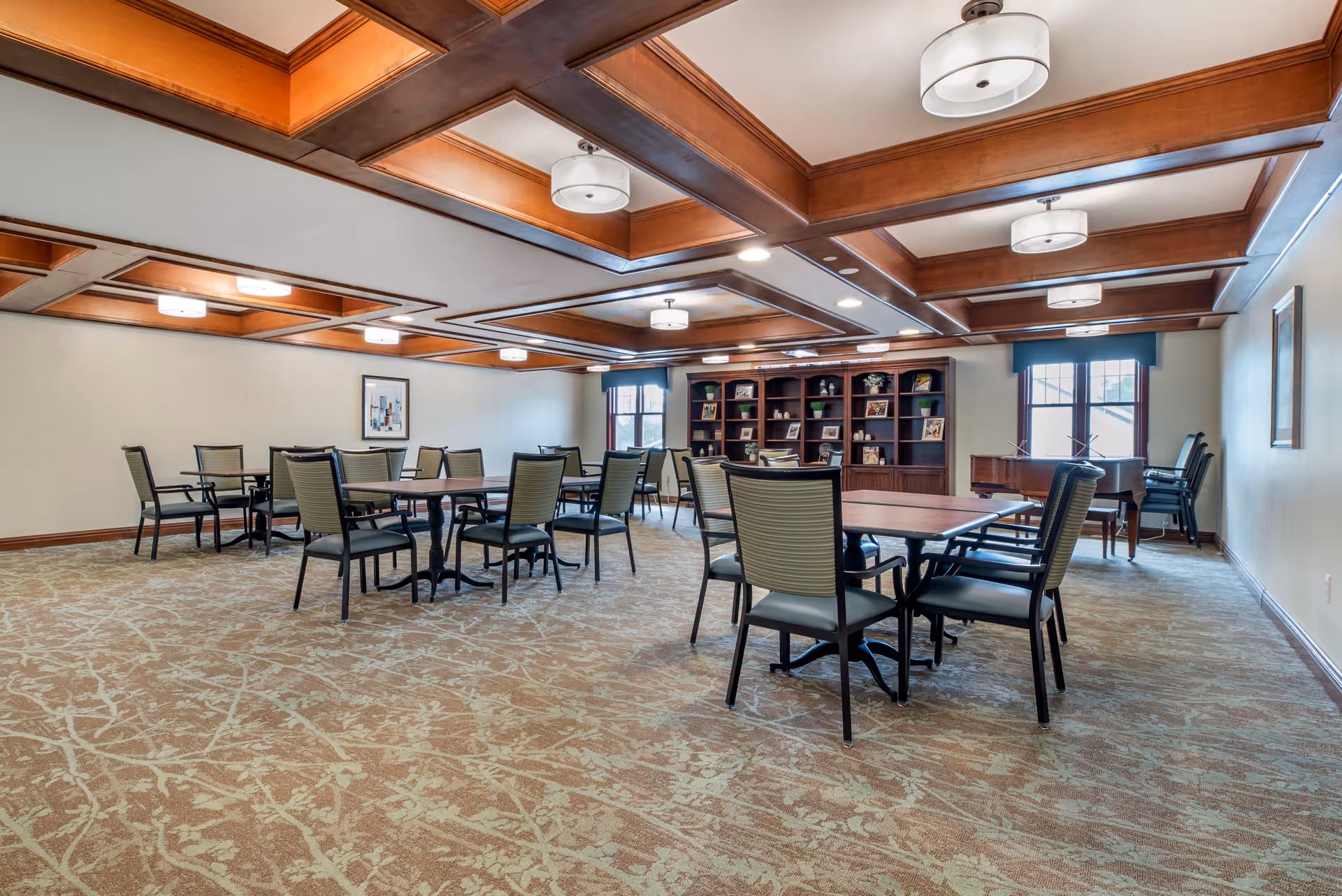 A spacious room with multiple tables and chairs arranged for group seating, featuring a patterned carpet, wooden coffered ceiling with modern light fixtures, large windows with blue valances, and a dark wooden bookshelf filled with decorative items along the back wall.