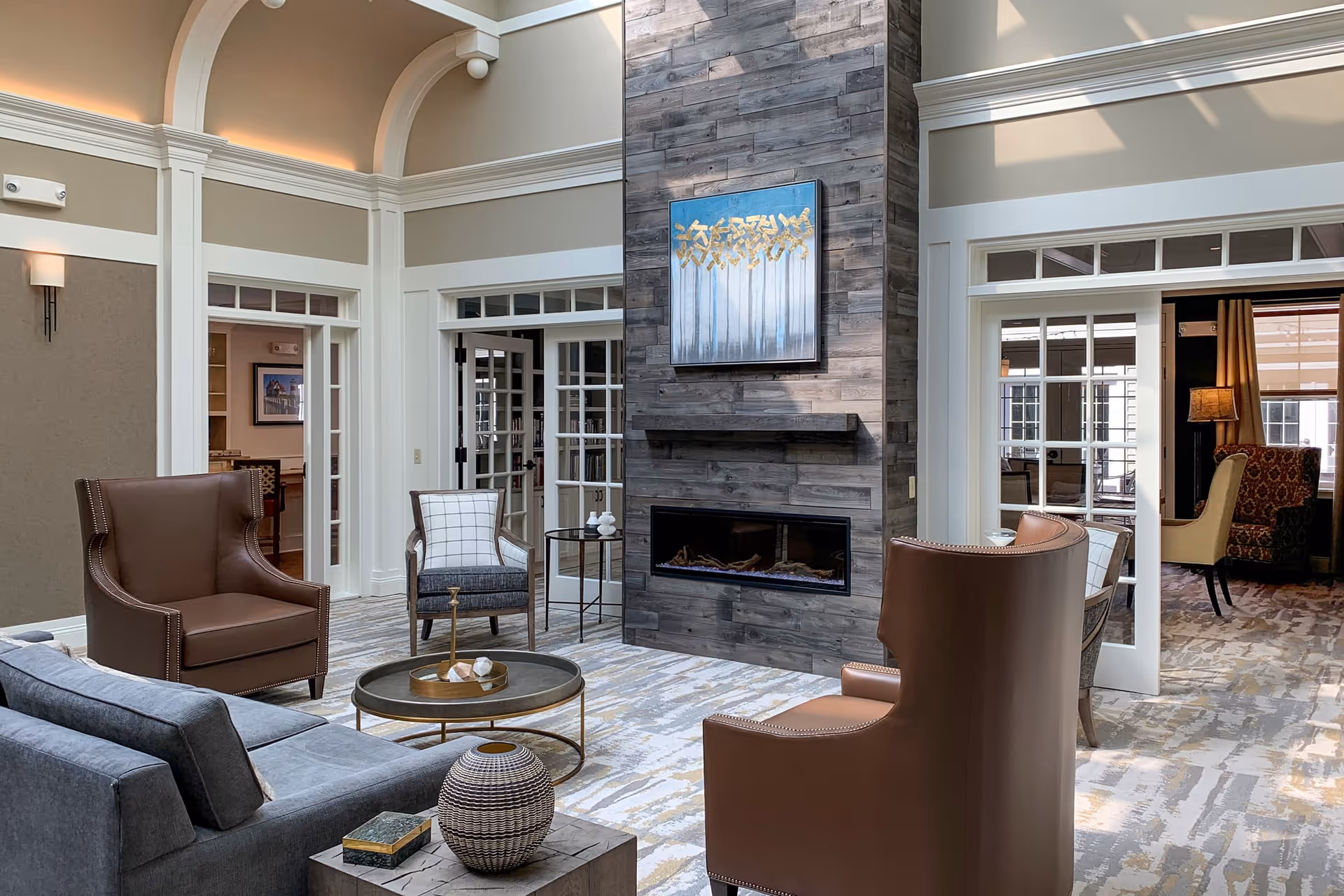A cozy living room area in a senior living facility featuring a modern fireplace with a wooden panel surround and a painting above it. The room has a high ceiling with soft lighting, a gray sofa, two brown leather armchairs, a patterned armchair, a round coffee table, and a decorative vase on a side table. French doors lead to adjacent rooms with additional seating and warm lighting.