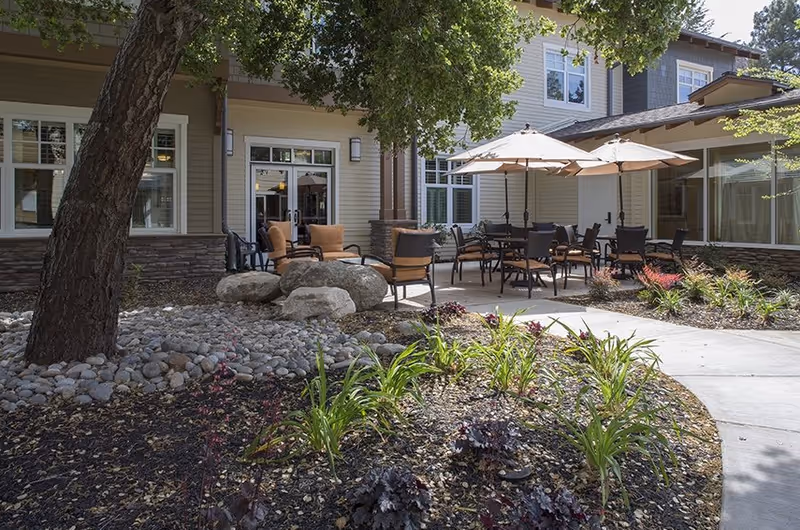 Outdoor patio area at The Terraces at Los Altos featuring several tables with umbrellas and chairs arranged for seating. The patio is surrounded by landscaping with plants, rocks, and trees, adjacent to a beige building with large windows and doors.