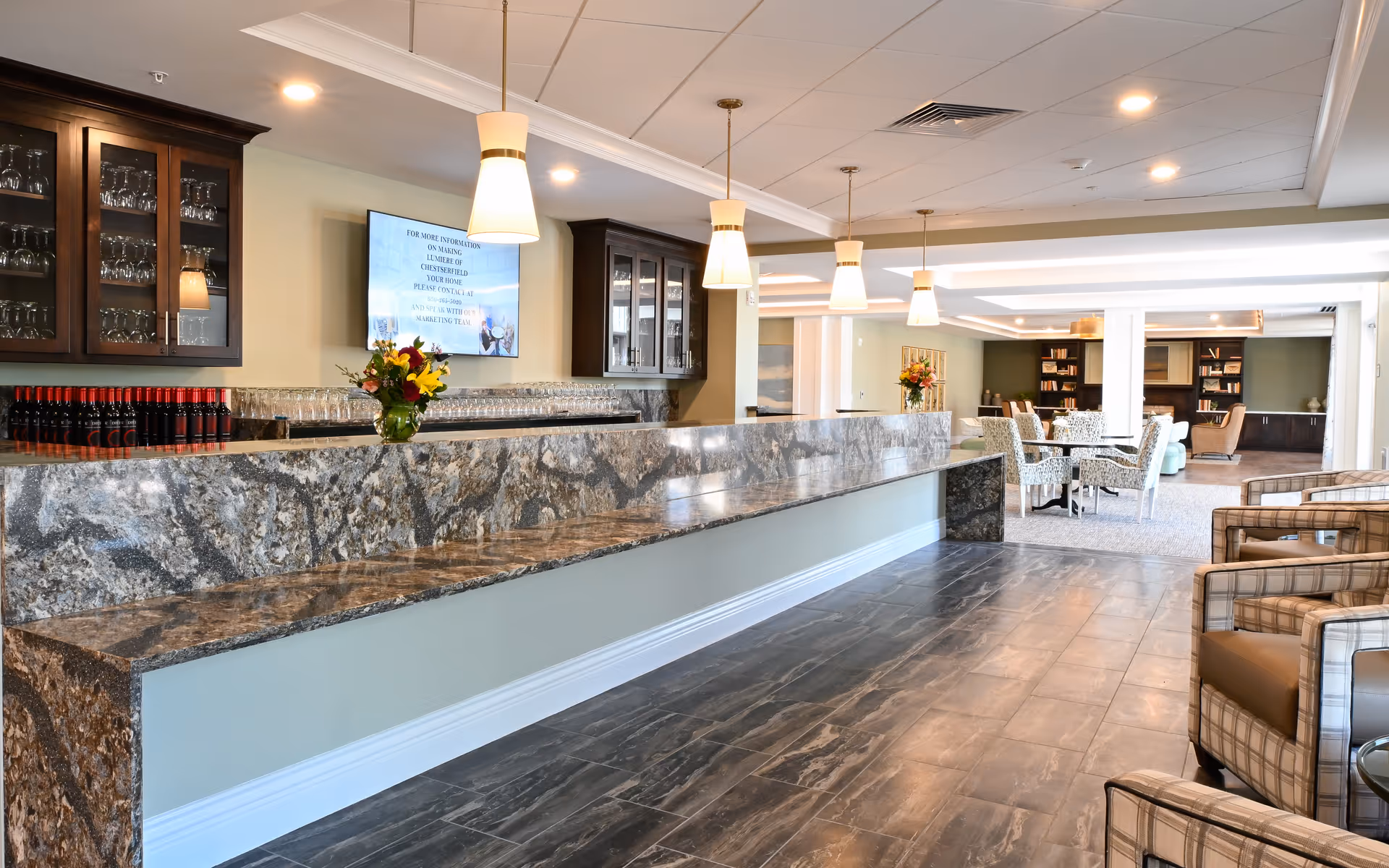 Spacious senior living lounge with a long marble-topped service bar, pendant lights, and seating areas with chairs and tables.