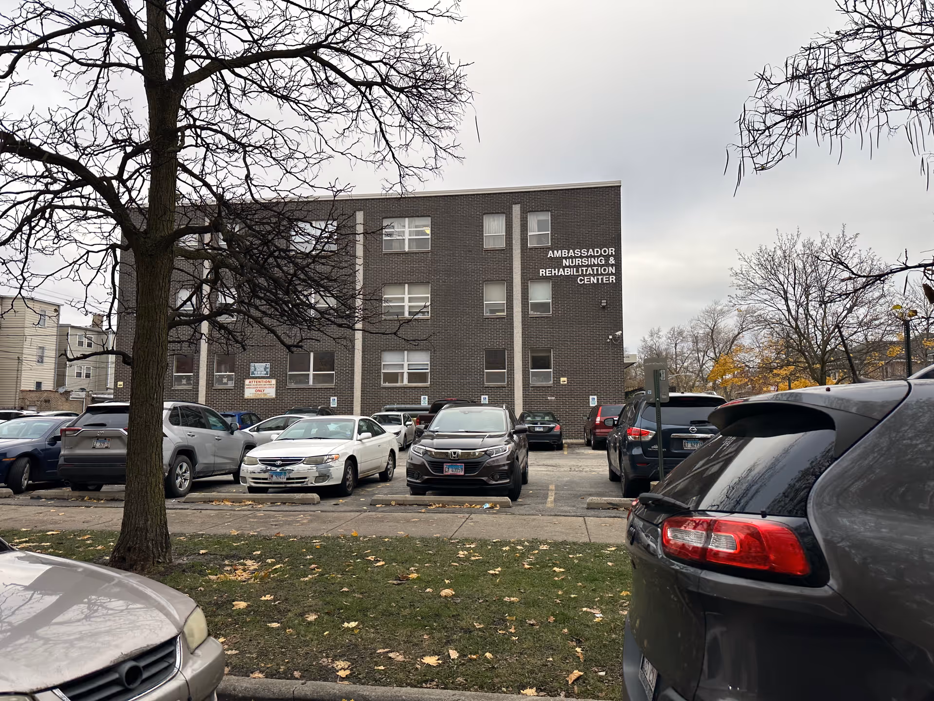 Exterior view of a three-story brick building with the sign 'Ambassador Nursing & Rehabilitation Center' on the upper right side. Several cars are parked in front of the building in a parking lot. Leafless trees and a cloudy sky are visible.