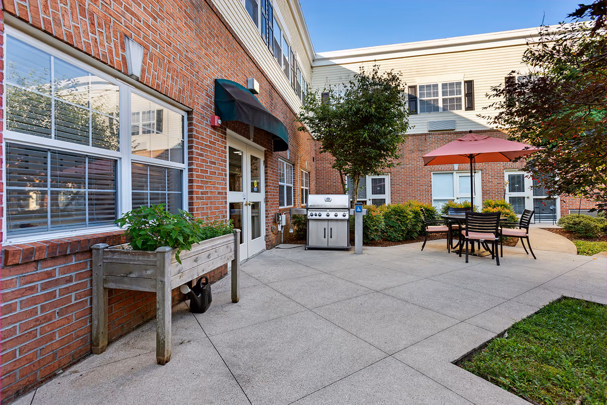 Courtyard patio beside a brick building featuring a grill, table with umbrella and chairs, planter box and entrance door.