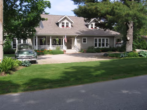 A single-story house with a large front porch, surrounded by trees and greenery. There is a vintage green truck parked on the left side of the driveway. The house has beige siding, white trim, and dormer windows on the roof. The lawn is well-maintained with a paved driveway leading to the porch.