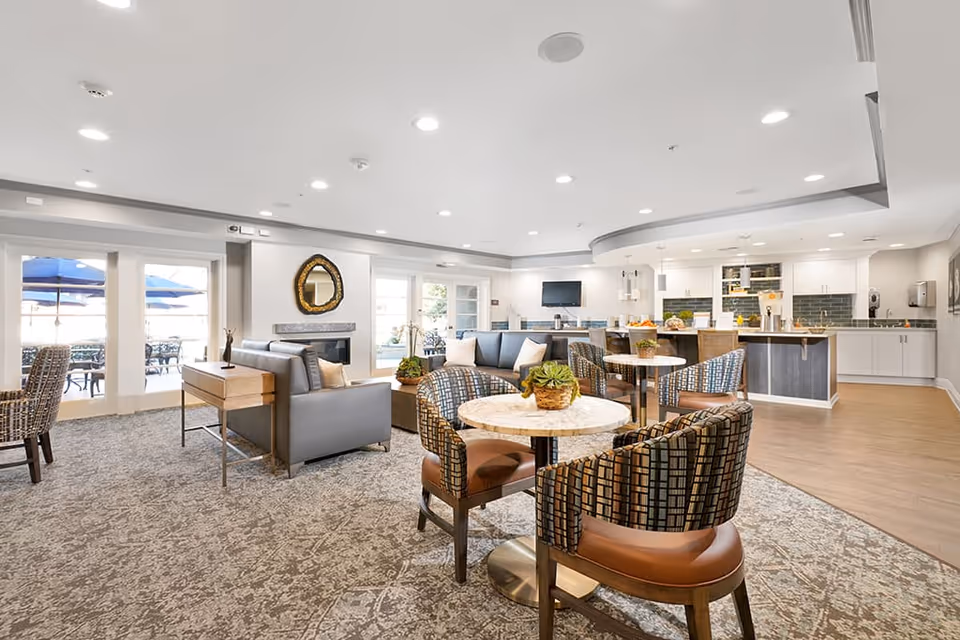 A bright and spacious common area in a senior living facility featuring multiple round tables with patterned chairs, a seating area with gray sofas and a decorative mirror above a fireplace, and a modern kitchen with white cabinets and a breakfast bar. Large windows and glass doors lead to an outdoor patio with blue umbrellas.