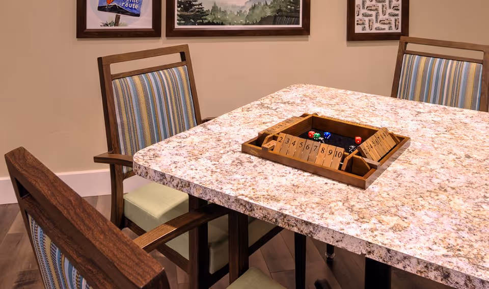 A close-up view of a square table with a granite-like surface and a wooden game set with dice in the center. Surrounding the table are wooden chairs with striped cushions. The wall behind has framed pictures.