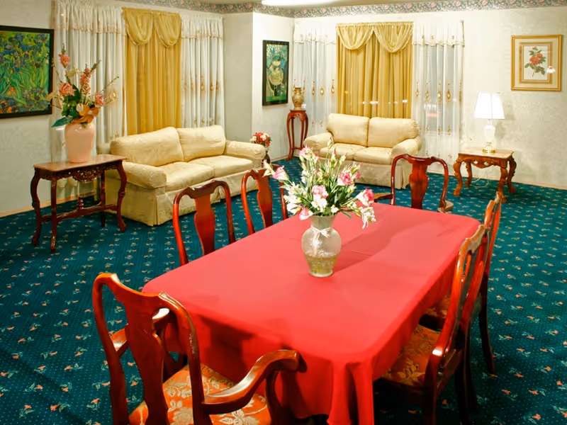 A cozy living room area with two beige sofas placed against walls with windows covered by yellow and white curtains. In the foreground, there is a dining table covered with a red tablecloth and surrounded by wooden chairs. A vase with flowers is placed on the dining table. The room has green carpet with a floral pattern, and there are side tables with lamps and decorative items along the walls.