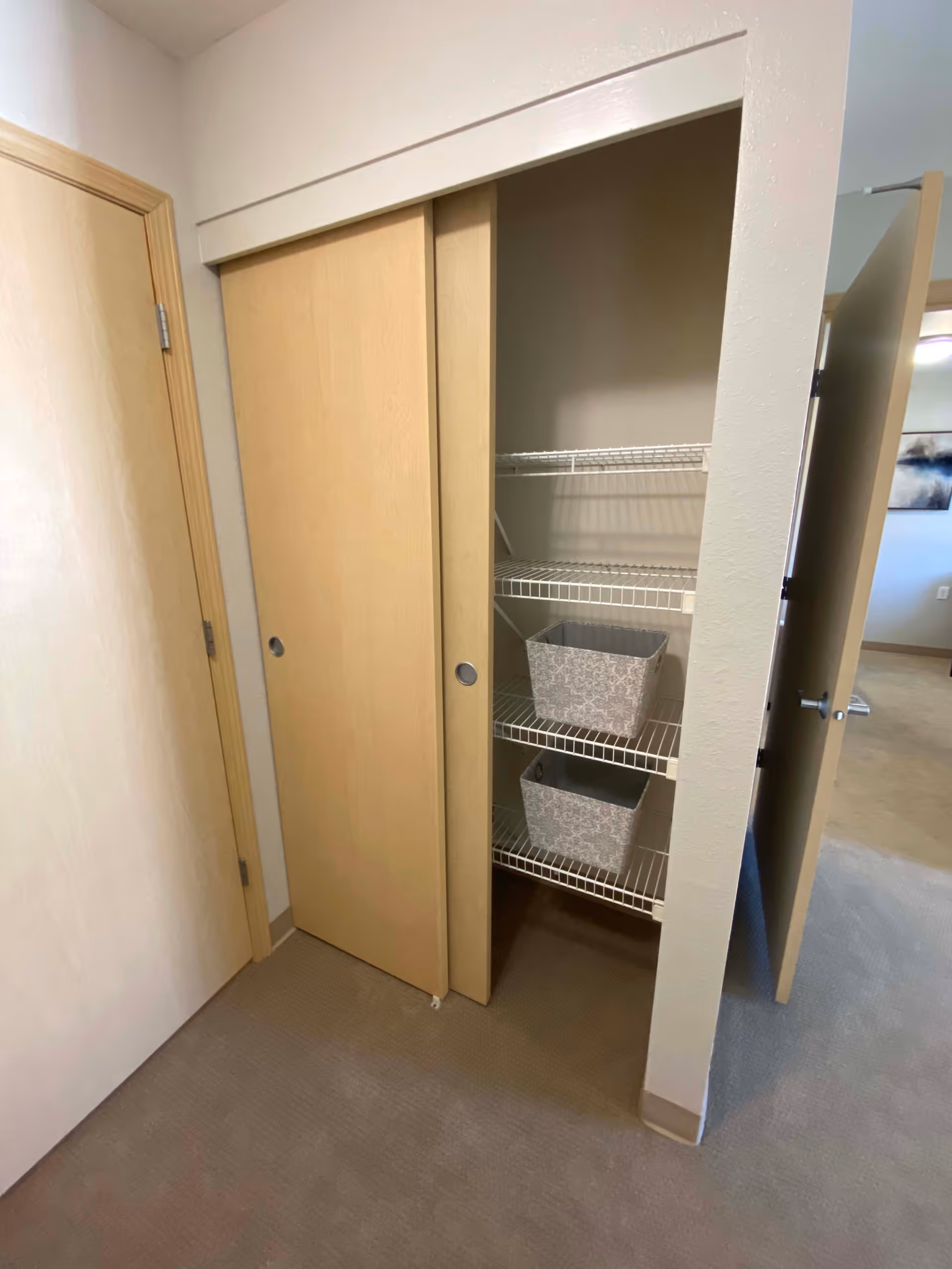 A small closet with sliding wooden doors, one door partially open revealing three white wire shelves inside. Two gray patterned storage bins are placed on the middle and bottom shelves. The closet is located in a carpeted room with beige walls and a wooden door to the left.