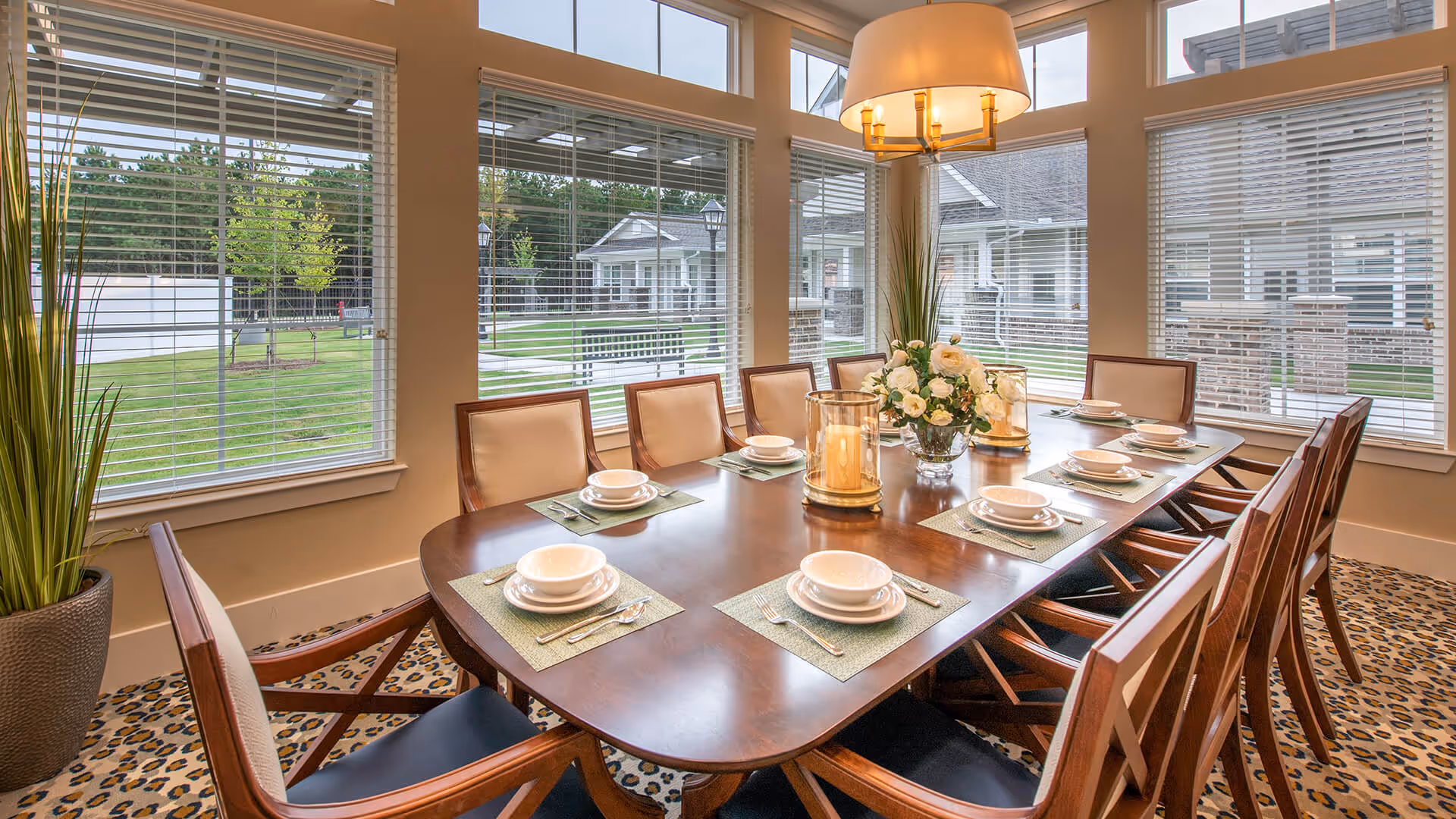 Sunlit dining room with a long wooden table set for ten, a floral centerpiece and candles, and large windows overlooking the outdoor grounds.