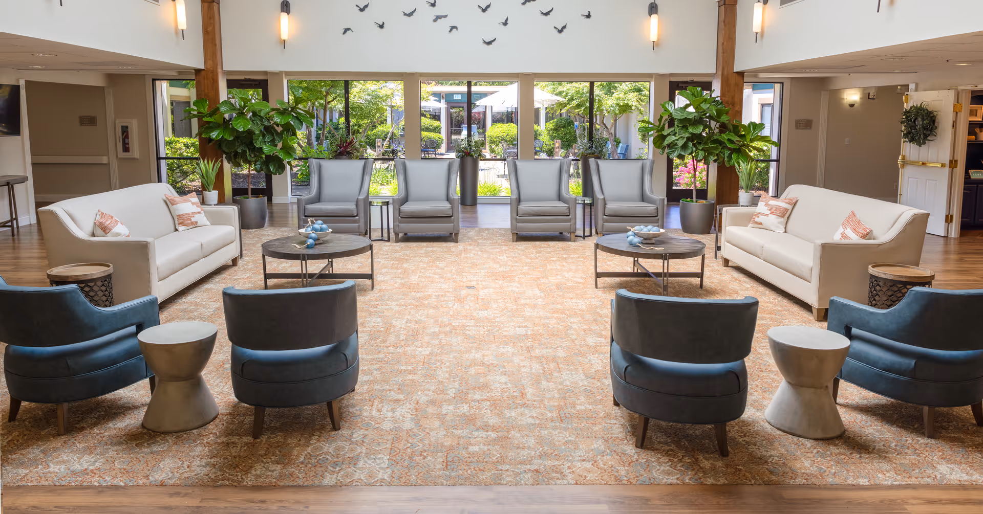 A spacious and well-lit senior living facility common area with two beige sofas, six gray armchairs, and four blue chairs arranged around two round coffee tables on a patterned carpet. Large windows at the back reveal a green outdoor garden area, and there are tall potted plants inside near the windows. The walls are decorated with bird silhouettes and wall sconces providing warm lighting.