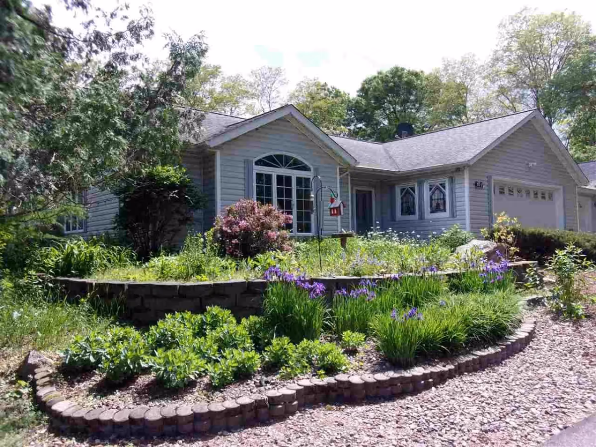 Single-story light-gray house with a landscaped front yard, flower beds, and a two-car garage.