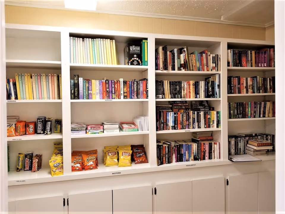 White built-in shelves filled with various books organized by genre including romance, mystery, westerns, and miscellaneous. Some shelves also contain snacks such as candy bars and chips. The shelves are labeled with genre names and snack categories.