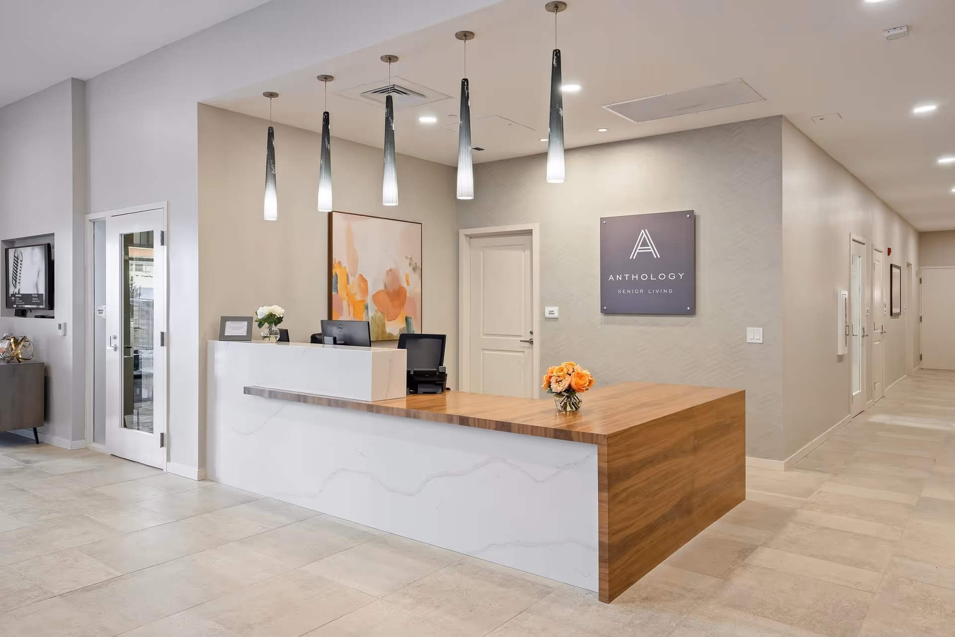 Reception area of a senior living facility with a modern front desk featuring a white marble and wood finish. Behind the desk are two office chairs, a computer, and a colorful abstract painting on the wall. Five pendant lights hang from the ceiling above the desk. A sign on the wall reads 'Anthology Senior Living.' The floor is tiled, and there is a hallway with several doors to the right.