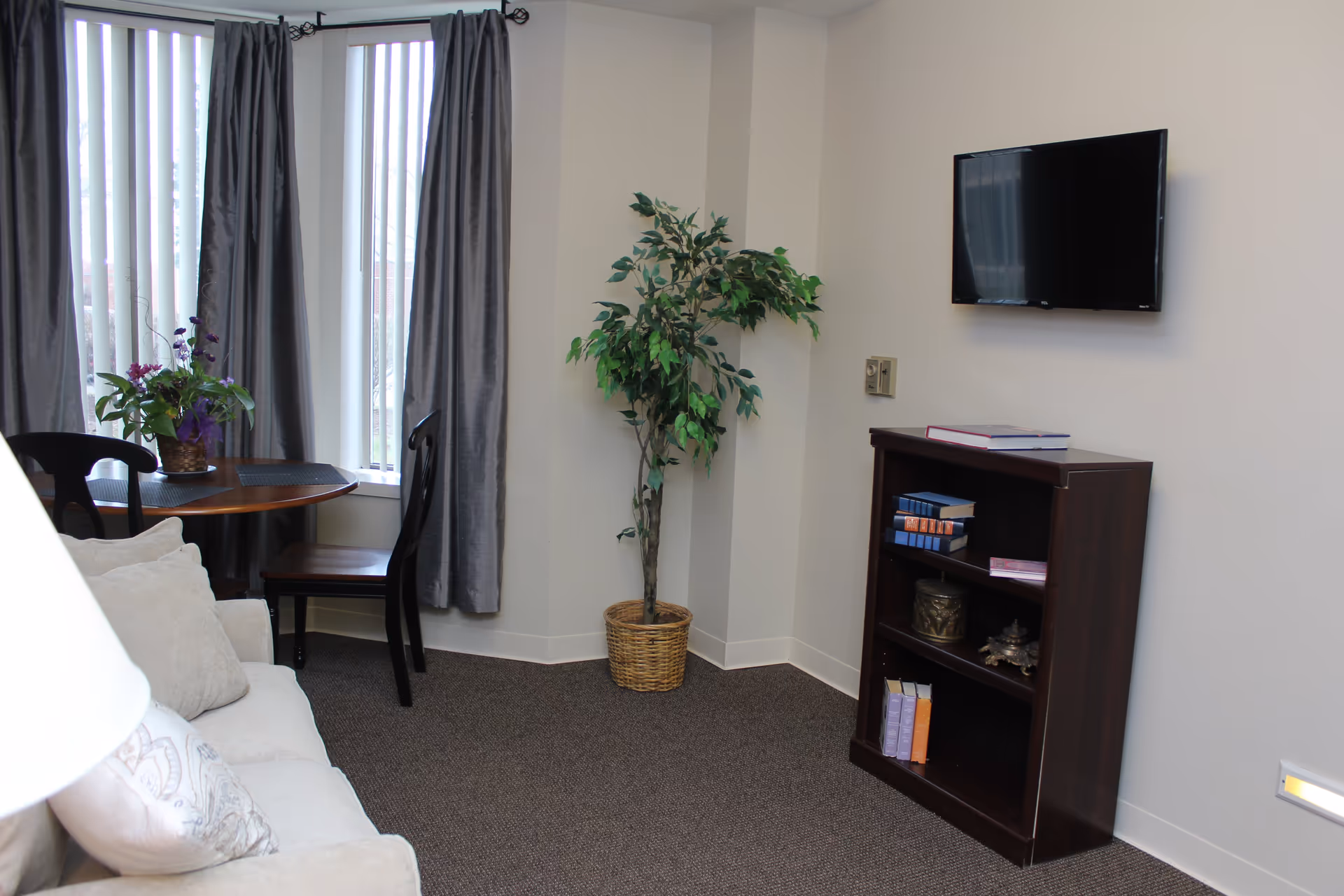 Small living room with a sofa, round table by tall windows, potted plant, wall-mounted TV, and a bookshelf.