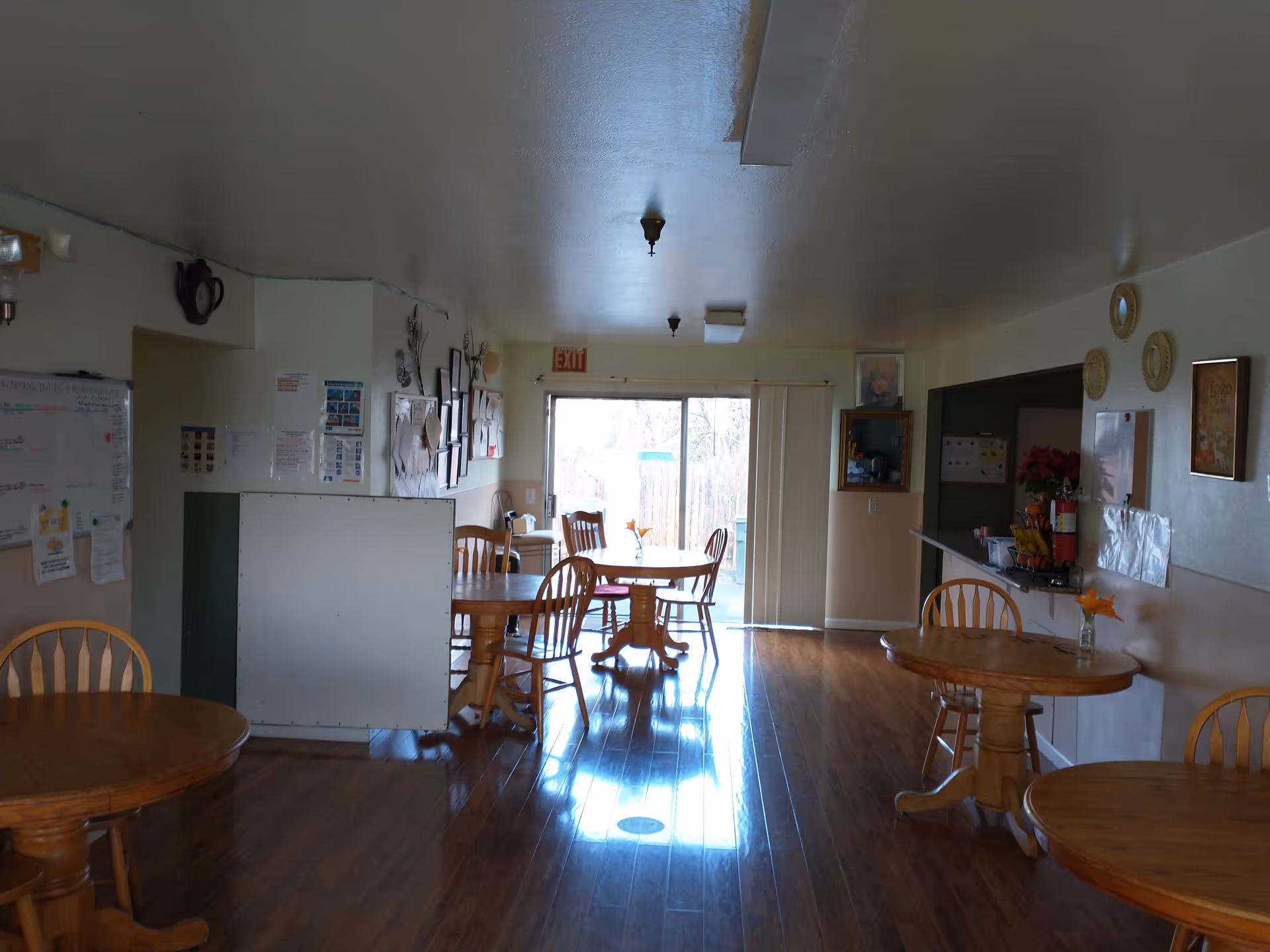 Interior view of a senior living facility dining area with wooden round tables and chairs arranged on a polished wooden floor. The room has light-colored walls decorated with framed pictures, a bulletin board with papers, and a sliding glass door at the far end letting in natural light. There is a counter area on the right side with a fire extinguisher and some fruit on display.