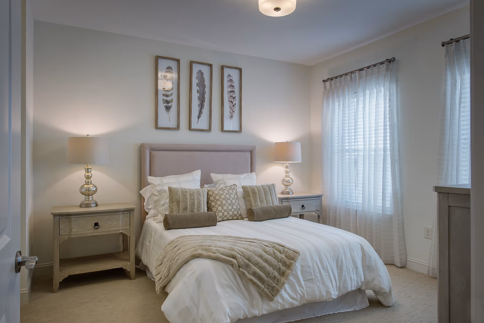 Well-lit bedroom with a neatly made bed, two bedside tables with lamps, framed feather artwork above the headboard, and sheer curtains over the windows.