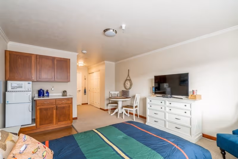 A cozy studio apartment featuring a bed with a colorful striped comforter in the foreground, a white dresser with a flat-screen TV on top, a small round dining table with two chairs, and a kitchenette area with wooden cabinets, a microwave, and a mini refrigerator. The walls are light-colored, and there is a decorative wall clock hanging above the dining table.