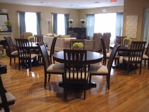 A spacious room with multiple round wooden tables and chairs arranged on a polished wooden floor. The room has several windows with blue curtains allowing natural light to enter. In the background, there are beige sofas and a piano, along with some decorative plants and flowers placed on tables and surfaces.