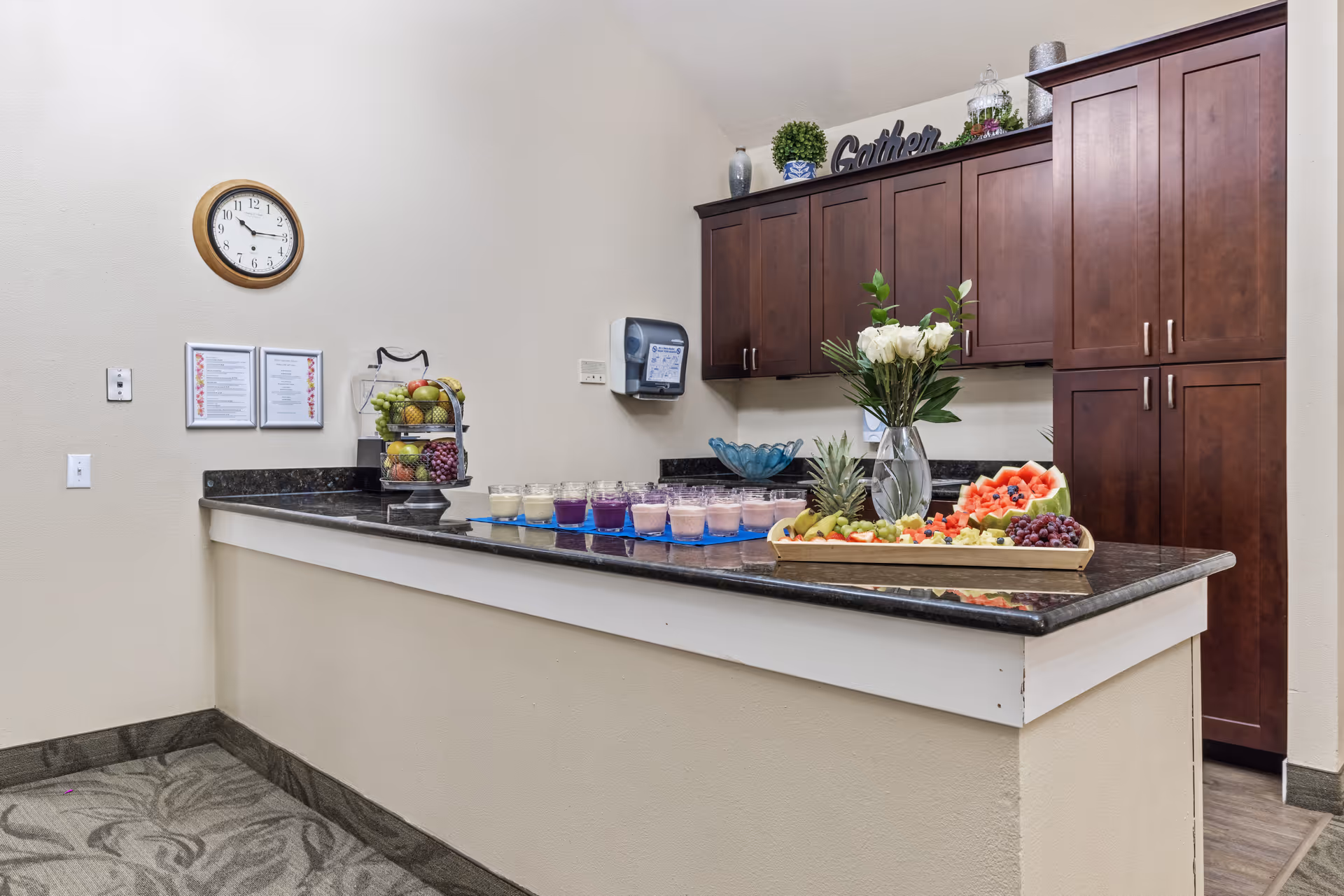 A kitchen area with dark wooden cabinets and a granite countertop. On the counter, there is a tray with assorted fresh fruit, a vase with white flowers, and several small cups filled with different colored smoothies or puddings. A clock and two framed notices are mounted on the wall, and a paper towel dispenser is attached near the cabinets. The word 'Gather' is displayed as decor on top of the cabinets.