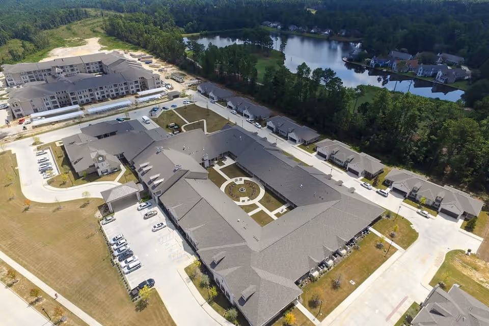 Aerial view of The Claiborne at Hattiesburg Assisted Living facility showing multiple buildings arranged around a central courtyard with a fountain, surrounded by parking areas, roads, and greenery, with a lake and residential houses in the background.