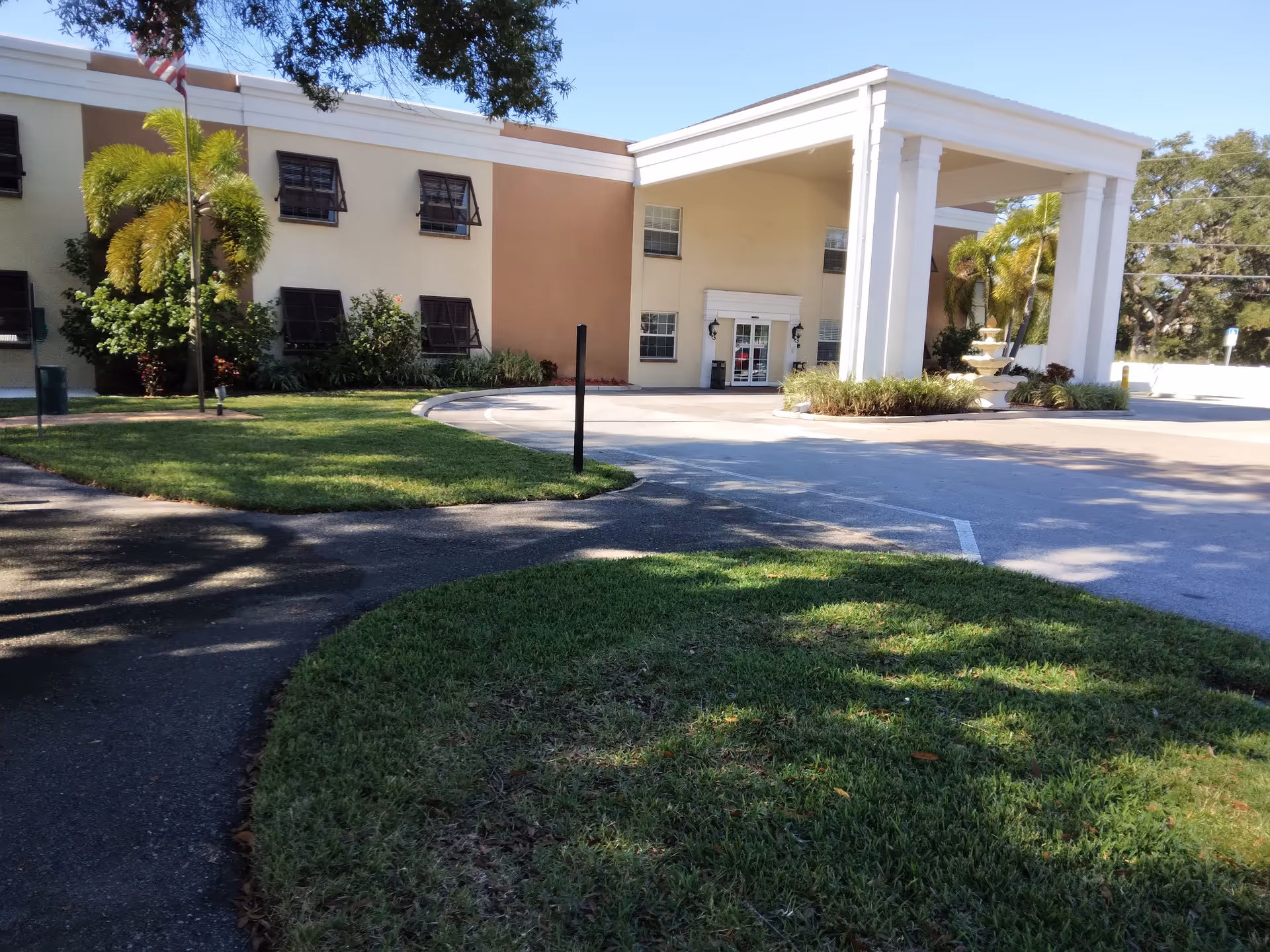 Exterior view of Volante Senior Living of Pinellas Park building entrance with a covered drop-off area supported by large white columns, surrounded by green grass, palm trees, and an American flag on a flagpole.