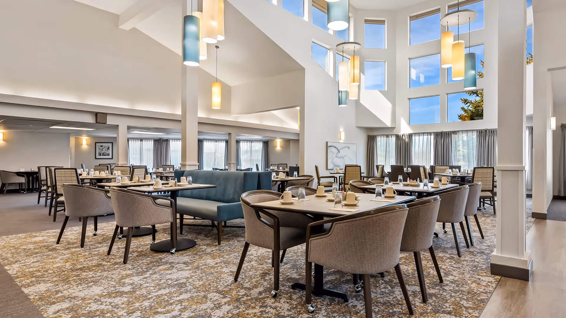 A spacious and well-lit dining room with multiple tables and chairs arranged neatly. The room features large windows allowing natural light to fill the space, modern pendant lights hanging from a high ceiling, and a patterned carpet covering the floor. Tables are set with cups, glasses, and napkins, ready for guests.