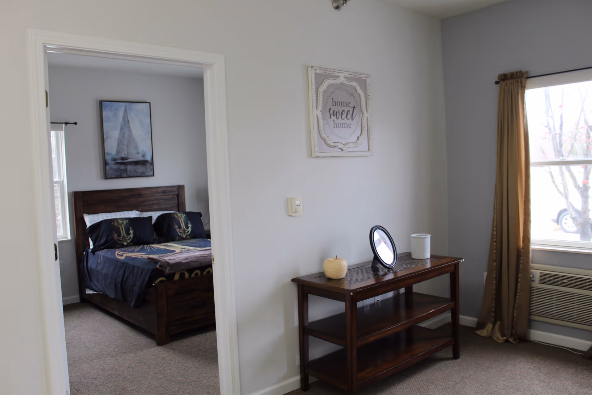 View of a cozy bedroom with a wooden bed frame, dark bedding featuring anchor designs, and a sailboat painting on the wall. Adjacent to the bedroom is a small area with a wooden console table holding a round mirror, a white candle, and a decorative pumpkin. A window with brown curtains and an air conditioning unit is visible on the right side.