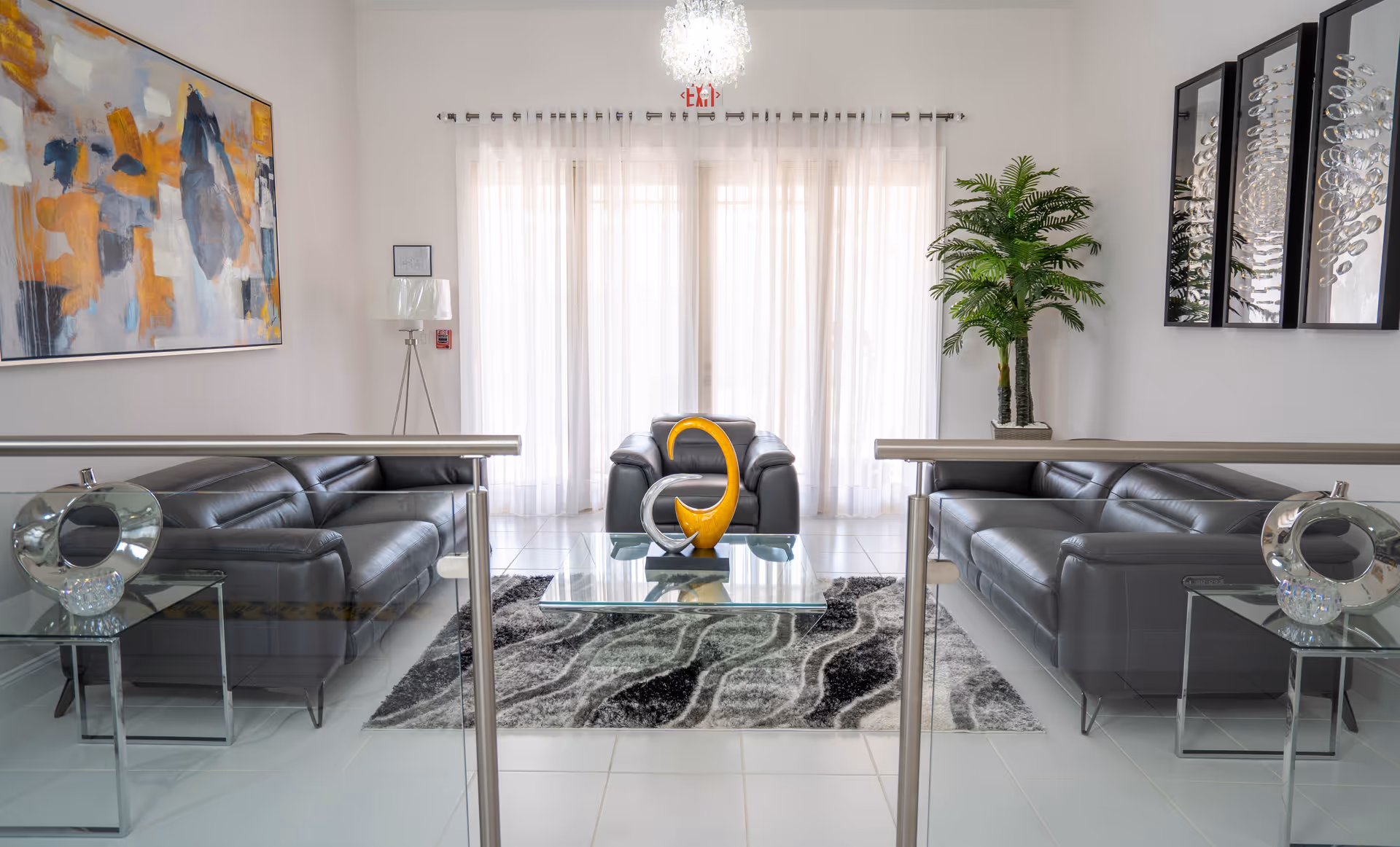 A modern living room with two black leather sofas facing each other and a matching armchair in the center. A glass coffee table with a decorative yellow and silver sculpture sits on a gray patterned rug. The room has white tiled floors, sheer white curtains covering large windows, a tall green plant in the corner, abstract wall art on the left, and three decorative mirrors on the right wall.